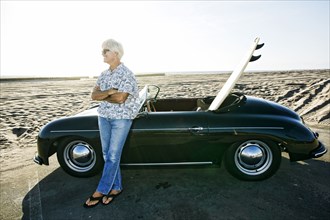 Older Caucasian man leaning on convertible car with surfboard on beach