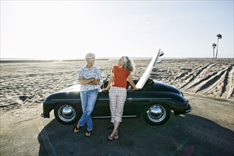 Older Caucasian couple leaning on convertible car with surfboard on beach