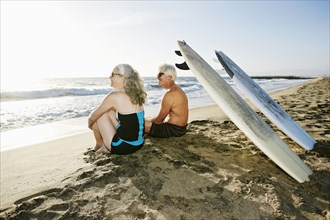 Older Caucasian couple sitting on beach with surfboards
