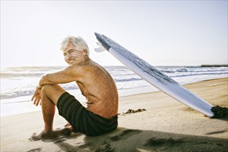 Older Caucasian man sitting on beach with surfboard