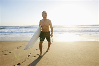 Older Caucasian man walking on beach carrying surfboard