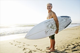 Older Caucasian man standing on beach carrying surfboard