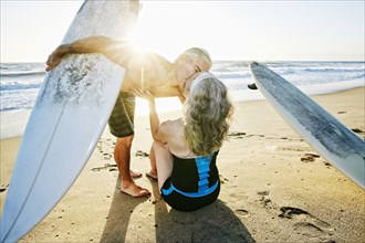 Older Caucasian couple on beach with surfboards kissing