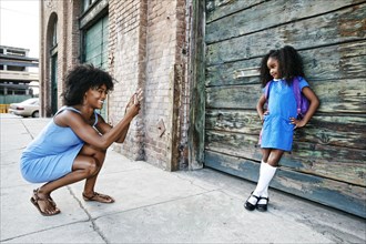Black mother photographing daughter on sidewalk with cell phone