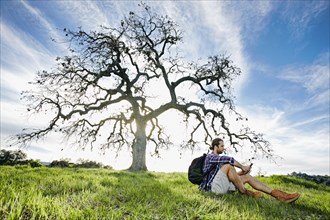 Caucasian man sitting in field near tree texting on cell phone