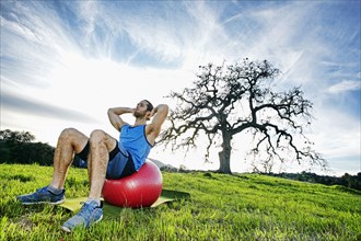 Caucasian man doing sit-ups on fitness ball in field near tree