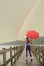 Caucasian woman in sportswear with red umbrella on pier catching rainbow