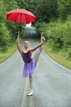 Caucasian ballerina dancing on remote road with umbrella