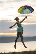 Caucasian ballerina jumping with multicolor umbrella on beach