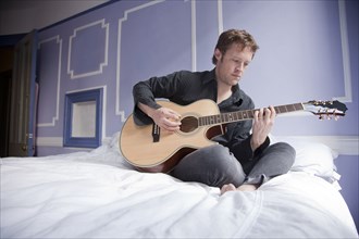 Caucasian man sitting on bed playing guitar
