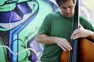 Caucasian man playing double bass in front of graffiti covered wall