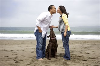 Husband kissing pregnant wife on beach while dog watches