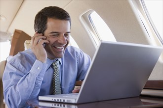 Hispanic businessman using laptop and cell phone on airplane