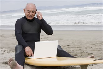 Senior Hispanic man sitting on surfboard using laptop and talking on cell phone