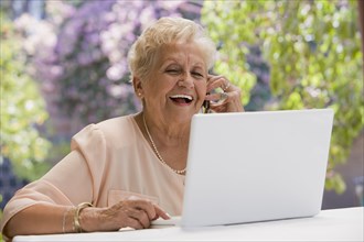 Senior Hispanic woman using laptop and talking on cell phone