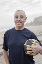 Hispanic senior man carrying soccer ball by beach