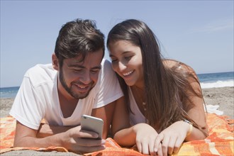 Hispanic couple laying on blanket at beach texting on cell phone