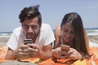 Hispanic couple laying on blanket at beach texting on cell phones