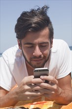 Hispanic man laying on blanket at beach texting on cell phone