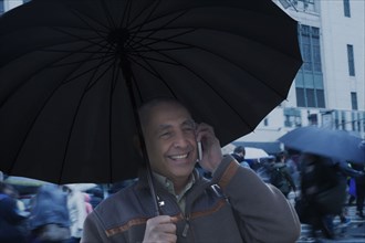 Smiling Hispanic man holding umbrella and talking on cell phone