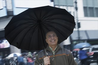 Smiling Hispanic man holding umbrella in city