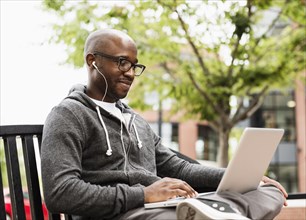 Black man using laptop and earphones on city bench