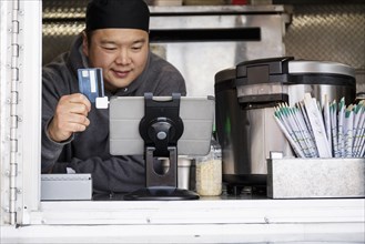 Asian man processing credit card using digital tablet at food cart