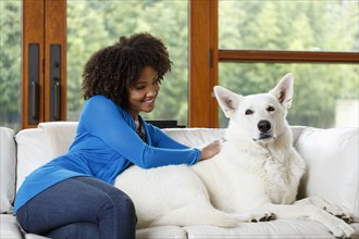 Smiling Black woman petting white dog on sofa