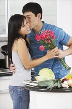 Asian man kissing girlfriend and giving her flowers in kitchen