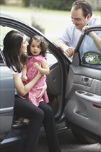 Hispanic mother and daughter sitting in car with door open talking to father