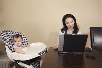 Chinese mother working on laptop next to baby in high chair
