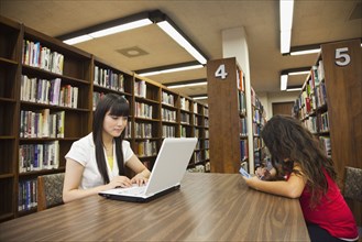 Woman on laptop and girl on video game in library