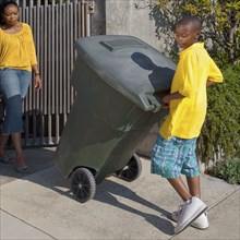 Black mother watching son wheel garbage can