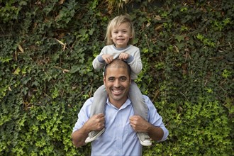 Hispanic father carrying son on shoulders near ivy wall