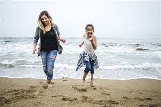 Hispanic mother and daughter walking barefoot on beach