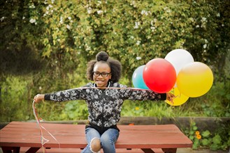 Black girl holding bunch of balloons on picnic table