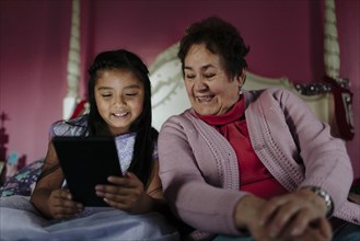 Hispanic grandmother and granddaughter using digital tablet on bed