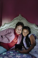 Hispanic grandmother and granddaughter using digital tablet on bed