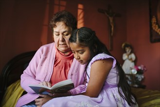 Hispanic grandmother and granddaughter reading book on bed