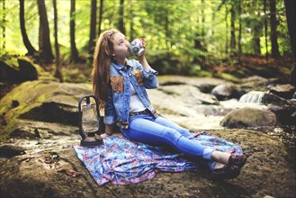 Caucasian teenage girl drinking coffee on forest rock