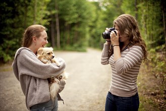 Girl photographing friend holding puppy in forest