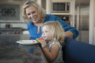 Grandmother watching granddaughter eating watermelon in kitchen