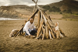 Father and baby son sitting under driftwood teepee on beach