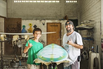 Father and son gesturing hang ten in surfboard workshop