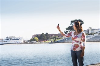 Hispanic woman taking cell phone selfie at waterfront