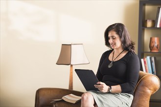 Hispanic woman sitting on armchair reading digital tablet