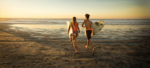 Teenage boy and girl carrying surfboards running to ocean