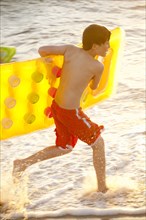 Caucasian boy running into ocean carrying inflatable raft