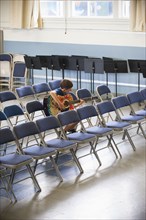 Caucasian boy sitting in row of chairs practicing guitar