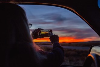Caucasian woman in car photographing sunset with cell phone